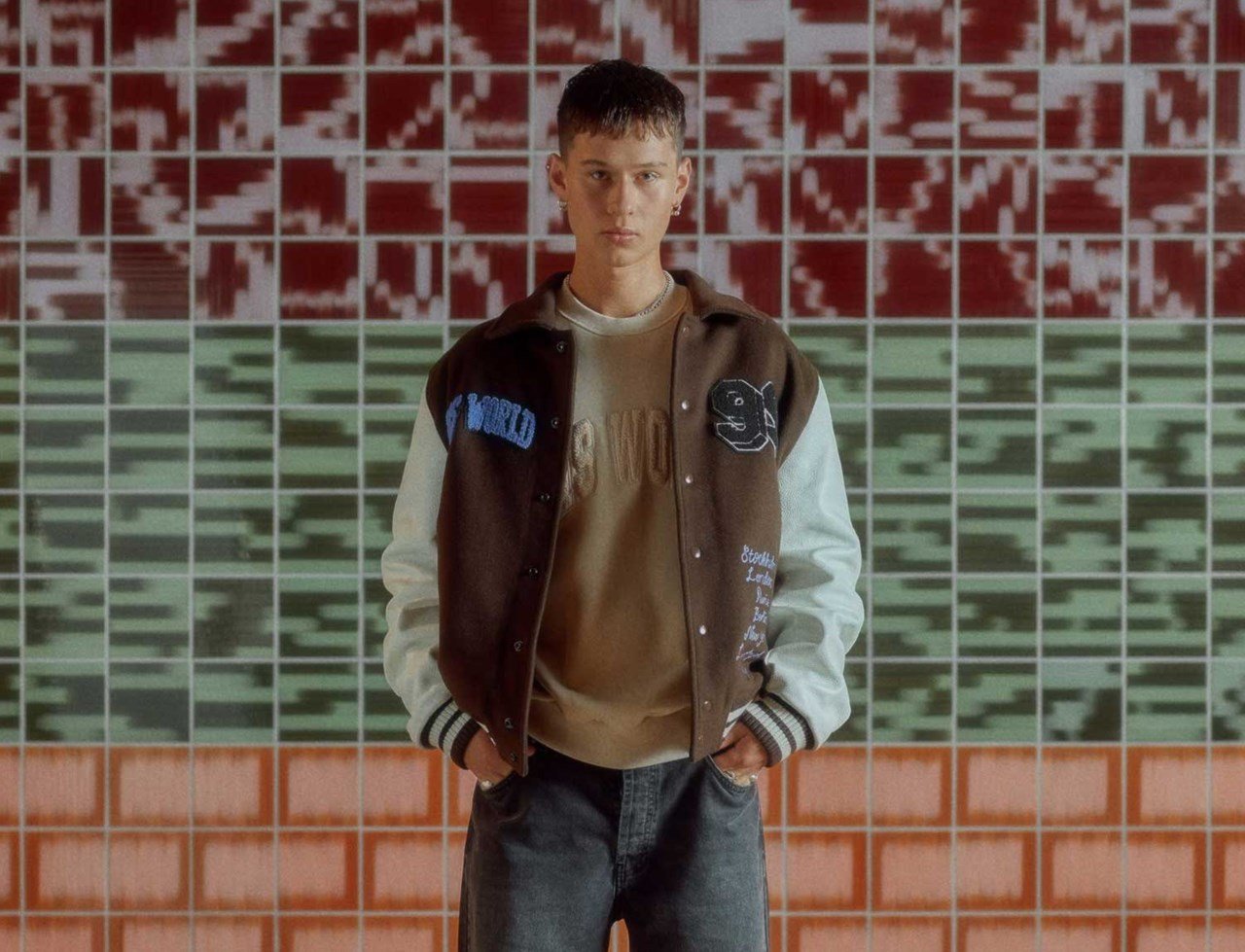 seasonal lookbook picture, man standing with a brown/white varsity jacket, beige crewneck and grey jeans